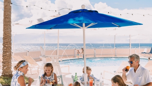 A family is sitting around a table with a beach and pool in the background.