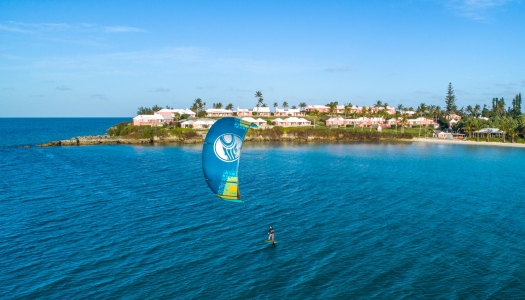 A person is kiteboarding on calm blue waters with a pink hotel in the background.