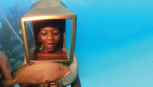 A young boy is wearing an underwater helmet looking at a fish.