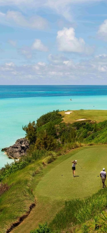 A wide angle of three people playing golf on a green grass with a various blue hued ocean in the background.