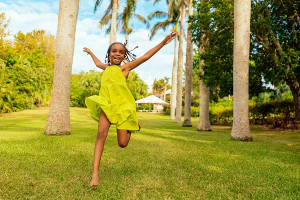 A smiling child in a green dress running barefoot through a patch of grass in Bermuda