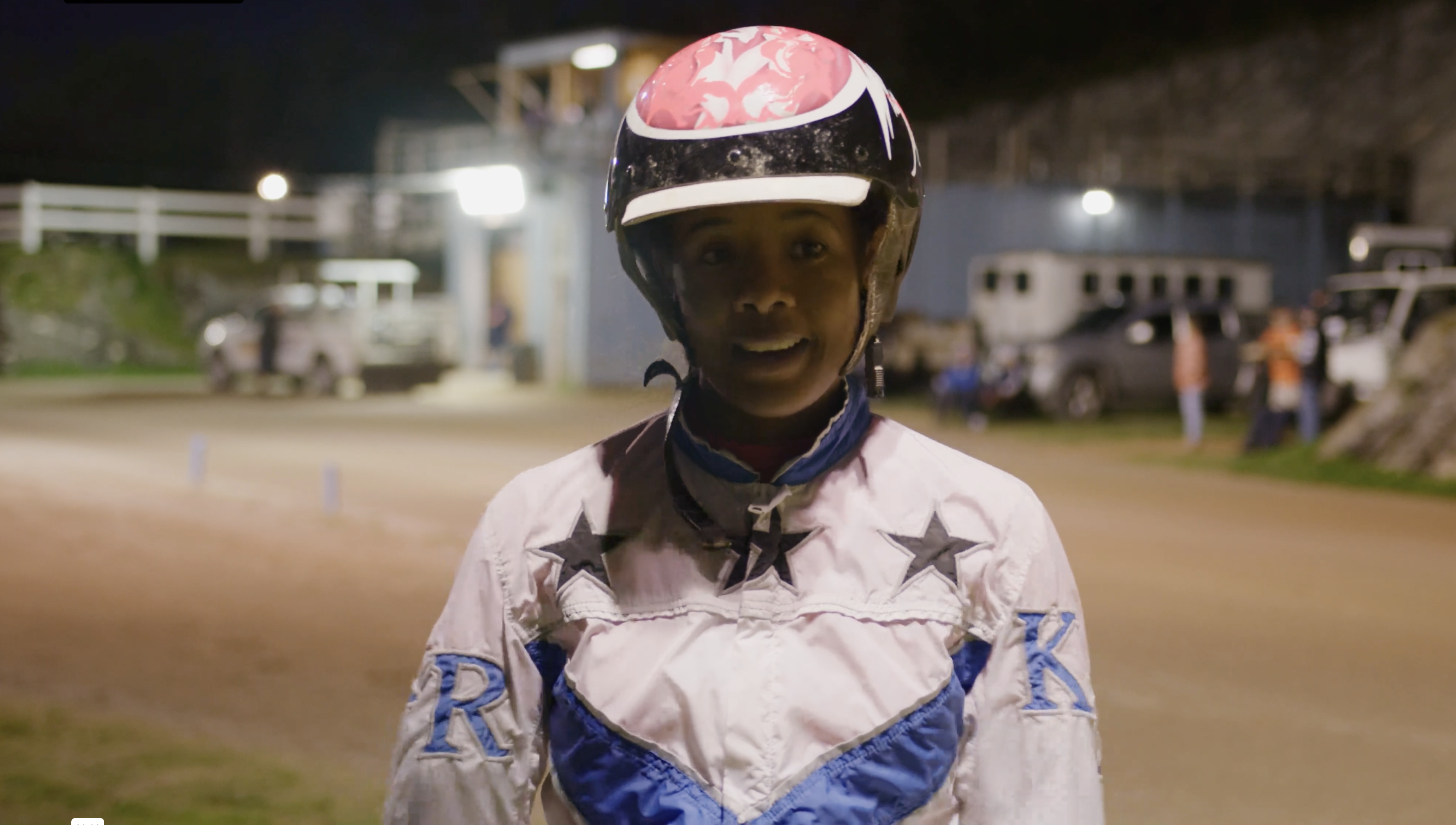 A jockey is talking to the camera with a horse track in the background.