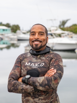A man is smiling in a wetsuit by a bay with boats behind him.