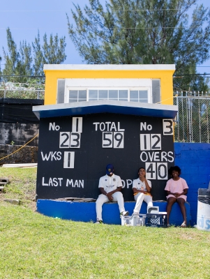 Three kids are sitting under the scoreboard at a cricket game.