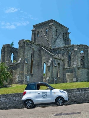 A person is driving an electric microcar by a scenic building.