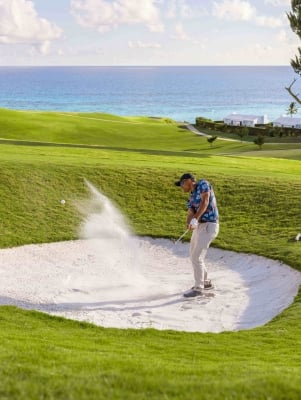 A man is hitting a golf ball out of a sand dune with the ocean in the background.