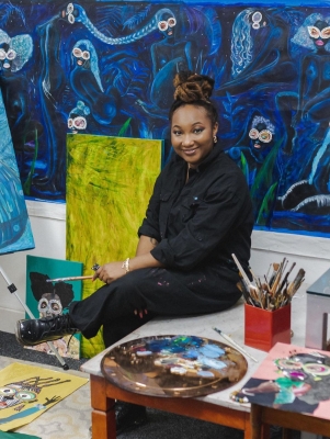 Gherdai Hassell is sitting on the floor surrounded by paintings and art supplies.