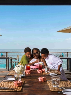 A woman and two children are celebrating a birthday a table with an ocean view in the background.