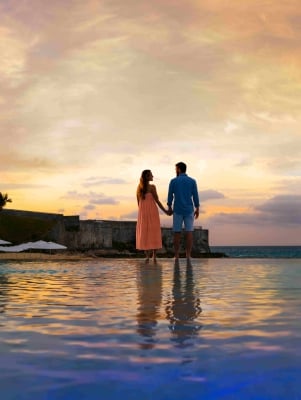 A couple is standing by a pool with sunset in the background.