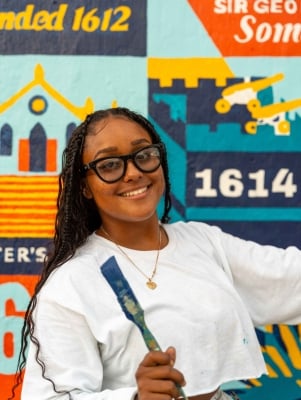 A woman is smiling at the camera with a paint brush.
