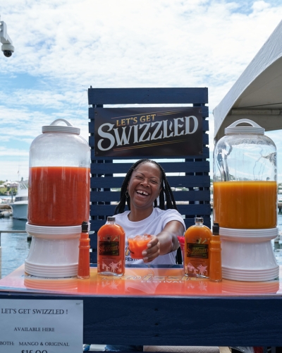 A woman is standing at a bar handing to the camera a rum swizzle. 