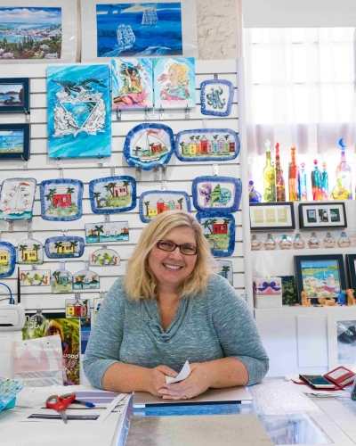 A woman is standing at the counter of her shop smiling at the camera.