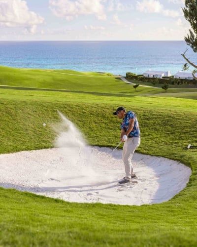 A man is hitting a golf ball out of a sand dune with the ocean in the background.