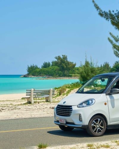 A woman is driving a micro car with a beach in the background.