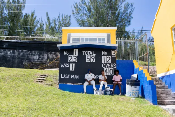 Three kids are sitting under the scoreboard at a cricket game.
