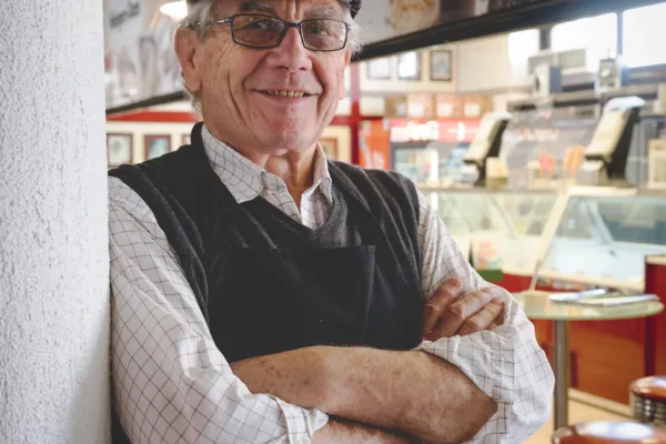 An older man is smiling at the camera in front of his shop. 