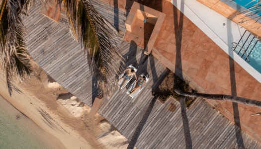 An aerial view of a couple sitting on lounge chairs with a beach in front and a snippet of a pool.