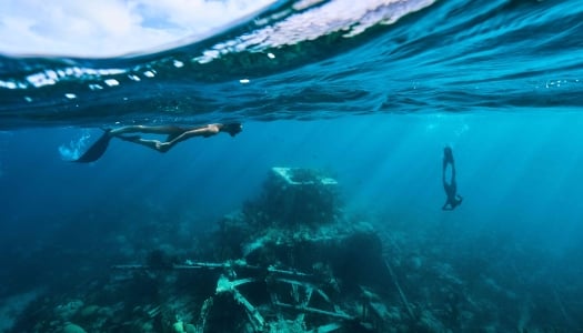 Two people are snorkeling in clear water around a shipwreck.