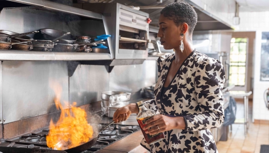 A woman is in a kitchen cooking in a pan with flames showing.