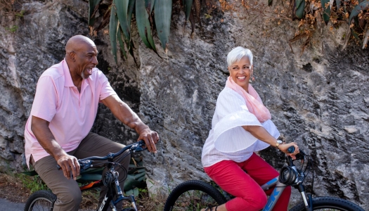 An older couple is riding bicycles and smiling at each other.