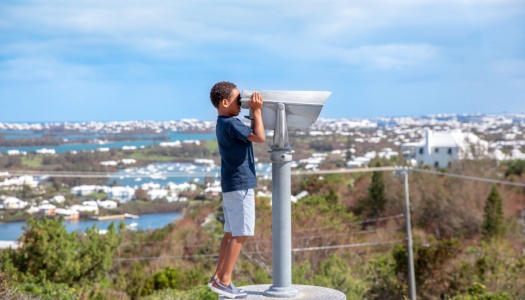 A little boy is looking through binoculars with a scenic background behind him.