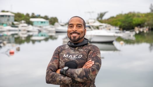 A man is smiling in a wetsuit by a bay with boats behind him.