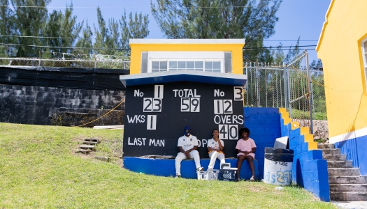 Three kids are sitting under the scoreboard at a cricket game.