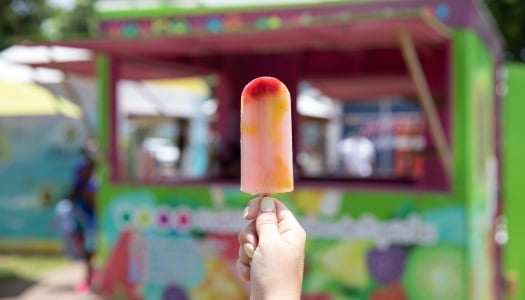 A hand is holding a ice pop with a colourful food stand in the background.