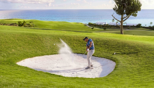 A man is hitting a golf ball out of a sand dune with the ocean in the background.