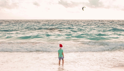 A little boy is standing on the beach in a Christmas hat.