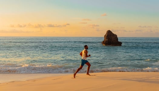 A man is running at sunrise on a empty beach with calm ocean waters in the background.