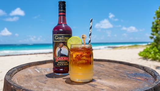 A view of a Goslings rum bottle with a dark n' stormy cocktail with a beach in the background.