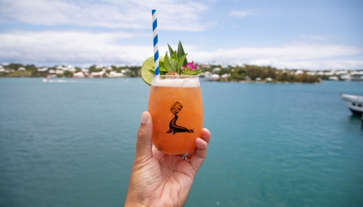 A person is holding a swizzle against a ocean backdrop.