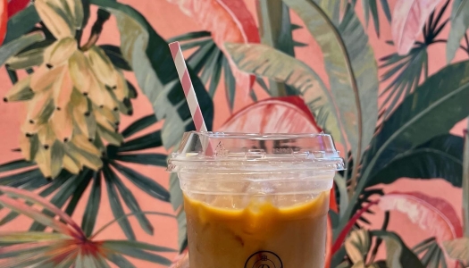 A hand holding up an iced coffee in front of tropical wallpaper