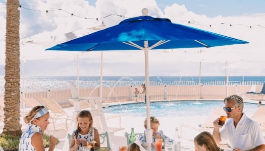 A family is sitting around a table with a beach and pool in the background.