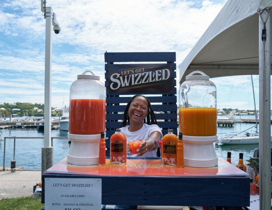 A woman is standing at a bar handing to the camera a rum swizzle.