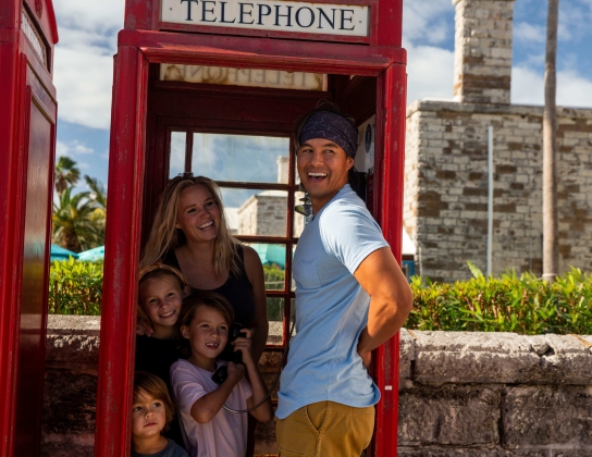 A family is piled up in an old style British telephone booth smiling at something in the distance.