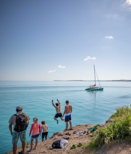 boat in the distance with people jumping off of a rock