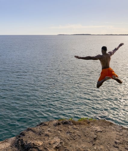 A man in orange shorts star jumps off of a cliff with calm ocean in the background.