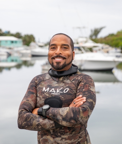 A man is smiling in a wetsuit by a bay with boats behind him.