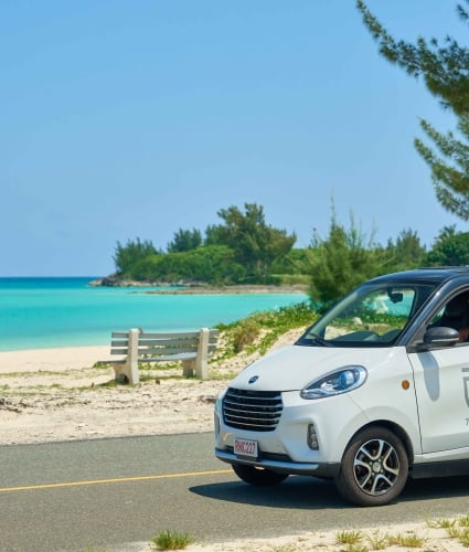 A woman is driving a micro car with a beach in the background.