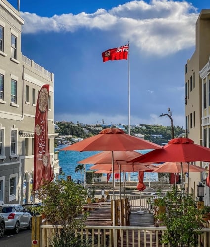 A restaurant deck with red umbrellas and a flag of Bermuda in the background.