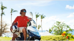 A man sits on a scooter with palm trees in the background.
