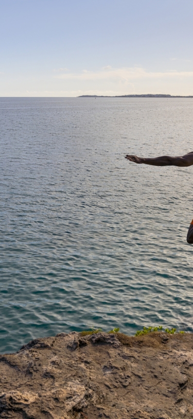 A man in orange shorts star jumps off of a cliff with calm ocean in the background.