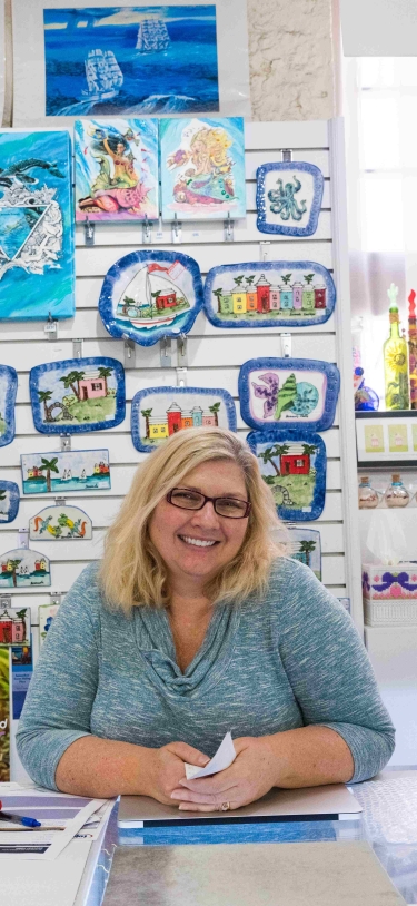A woman is standing at the counter of her shop smiling at the camera.