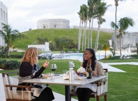 Two women are sitting eating dinner with a fort in the background.