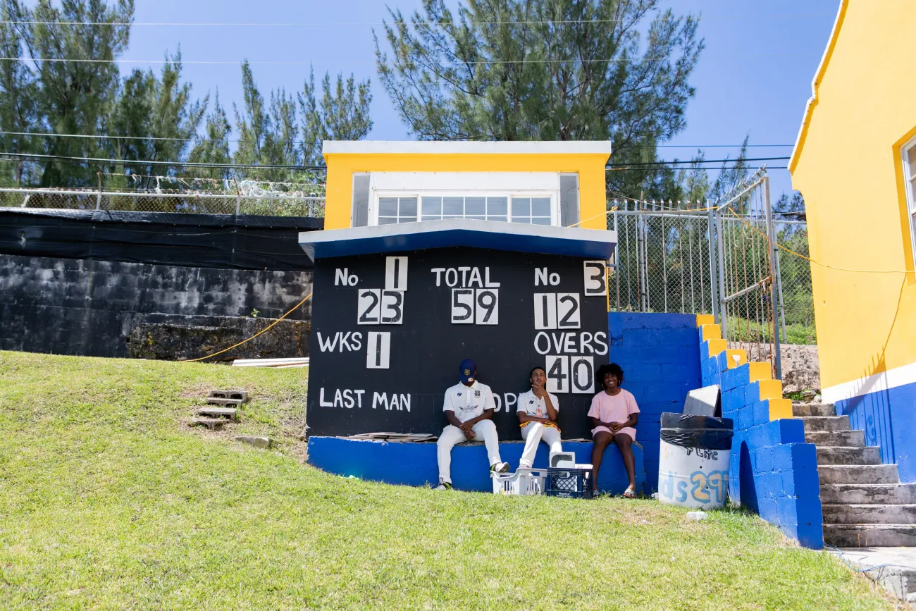 Three kids are sitting under the scoreboard at a cricket game.