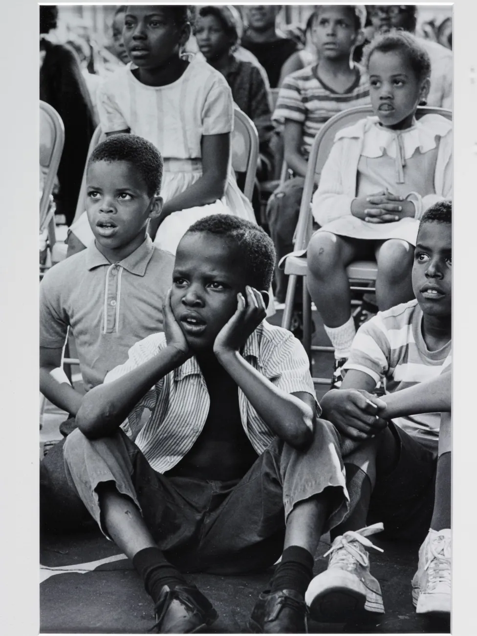 A group of young black children are watching a street theatre.