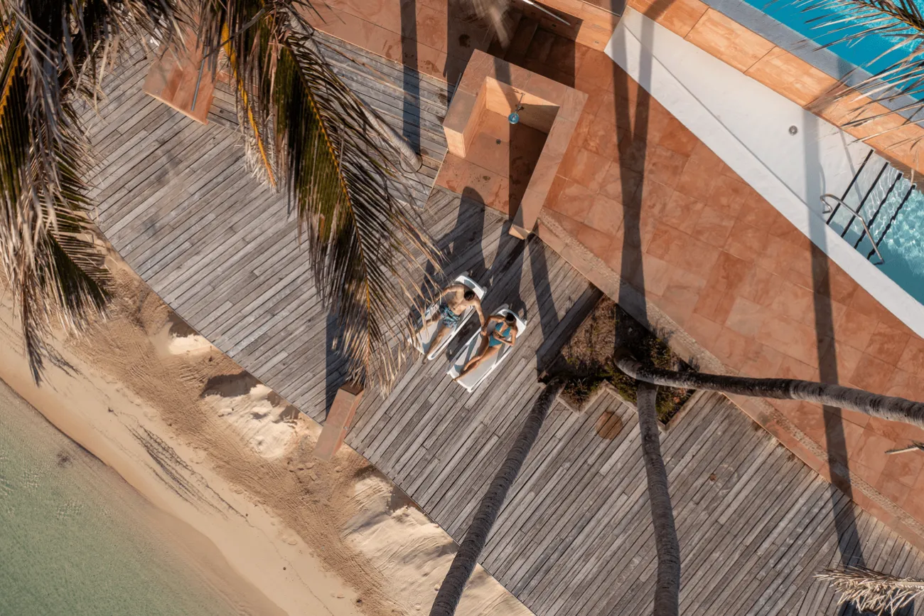 An aerial view of a couple sitting on lounge chairs with a beach in front and a snippet of a pool.