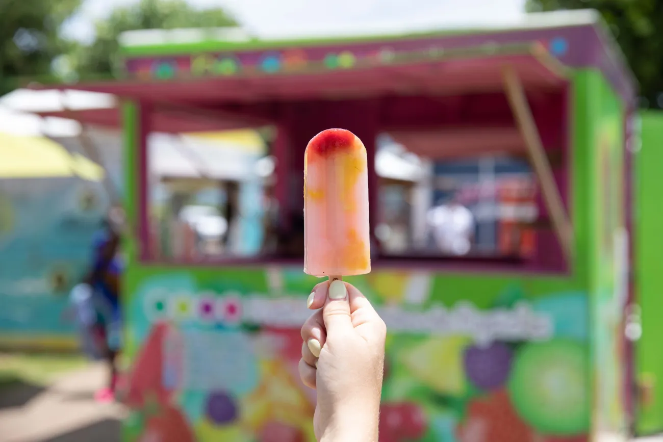 A hand is holding a ice pop with a colourful food stand in the background.